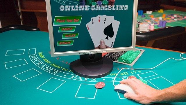 Hand på datormus vid datorskärm med bild på online gambling och en hand som håller fyra ess, allt detta på ett blackjackbord.